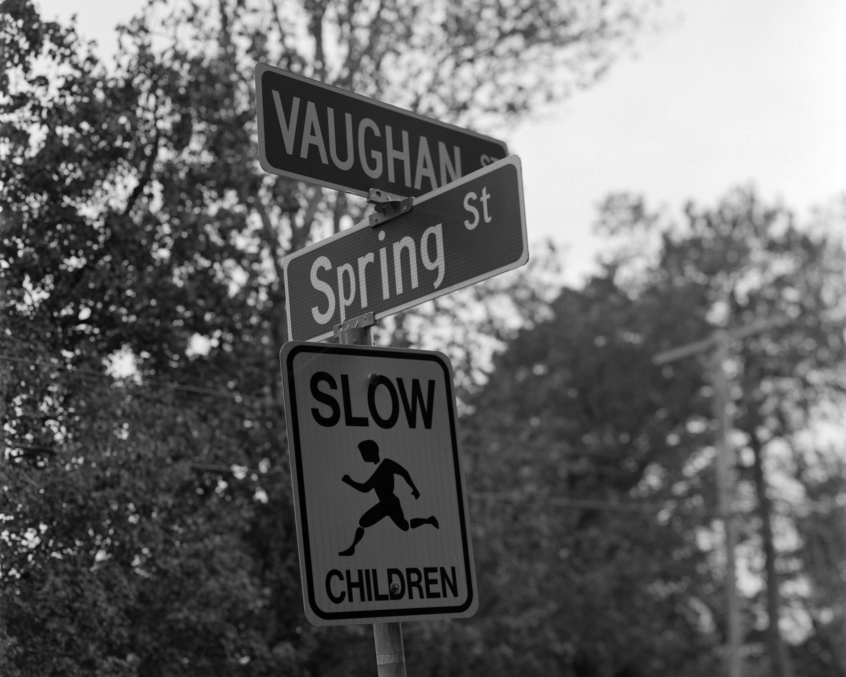 Black and white photo showing street signs at  intersection of Spring St and Vaughn St with Slow Children sign.